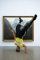 Nassim Baddag stands upside down in front of the painting by Carl Gustav Carus "Hochgebirge’. One hand is on the ground, the other is held vertically upwards. His legs are also stretched upwards. He is wearing black pants and a yellow T-shirt.