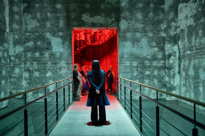 A dark corridor with concrete walls and a person with long hair looking at a red-lit opening.