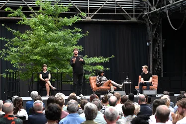 Jahrhunderthalle Bochum: Blick auf die Bühne der Veranstaltung "Brave New Voices". Es sind die Moderation Fatima Khan und der Schriftsteller Édouard Louis zu sehen. Daneben sieht man zwei Dolmetscher, die abwechseln in Deutsche Gebärdensprache übersetzen.  Im Vordergrund sieht man das Publikum.