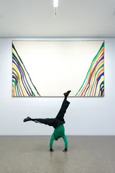 Nassim Baddag is standing on his head in front of the painting ‘Ksi’ by Morris Louis, 1960/61. He is wearing a green shirt and black pants. His hands are on the floor and his legs are at a right angle.