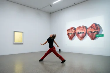 Nina Godderis wears a black shirt and red trousers. She dances in front of Yves Klein's work ‘MG 28’, 1960 and Oliviero Toscani's work ‘United Colors of Benetton / White Black Yellow, 1996’