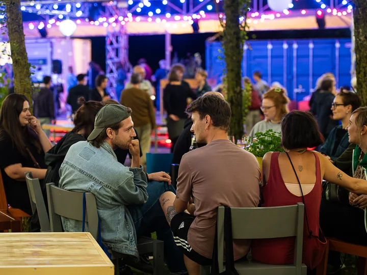 Junge Menschen sitzen bei Nacht in der Pappelwaldkantine der Ruhrtriennale, beleuchtet von Lichterketten