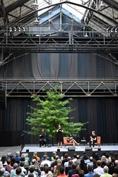 Jahrhunderthalle Bochum: Blick auf die Bühne der Veranstaltung "Brave New Voices". Es sind die Moderatorin Fatima Khan und der Schriftsteller Édouard Louis zu sehen. Daneben sieht man zwei Dolmetscher, die abwechseln in Deutsche Gebärdensprache übersetzen.  Im Vordergrund sieht man das Publikum.